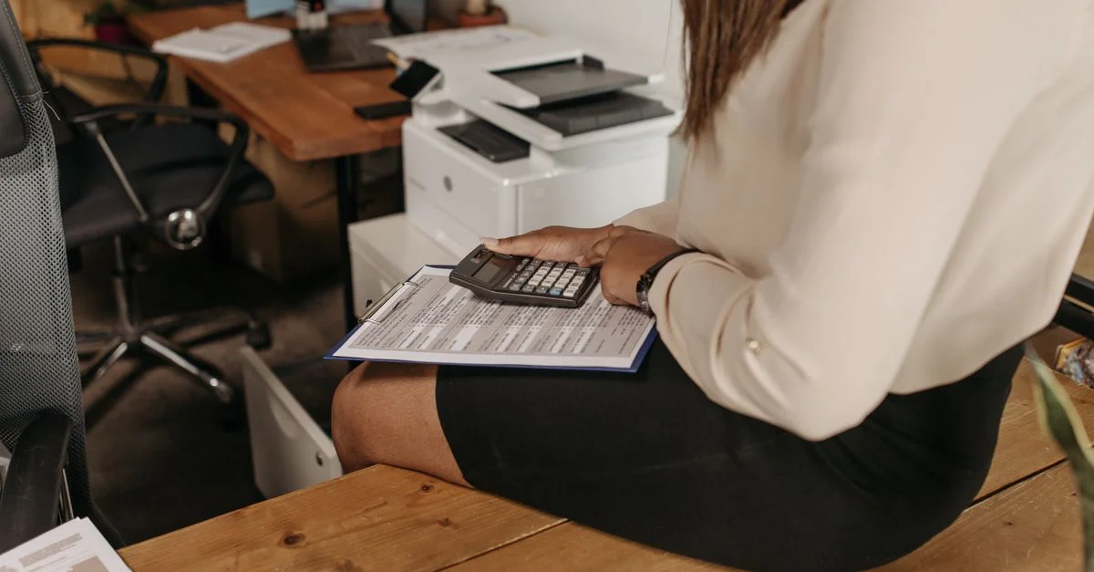 Une femme professionnelle est assise à son bureau en train de calculer ses finances avec un clipboard et une calculatrice.