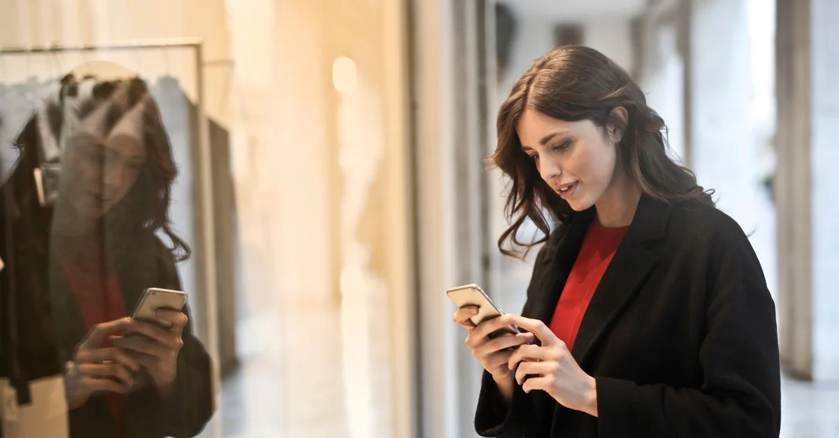 Jeune femme utilisant un smartphone devant une vitrine, reflétant son image, dans une tenue décontractée.